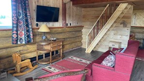 Ein Interieur einer Blockhütte mit einer Holztreppe, einem an der Wand montierten Fernseher, einem roten Sofa, einem hölzernen Schaukelpferd und einem gemusterten Teppich.