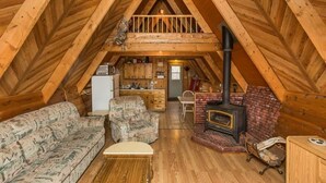 A cozy attic bedroom with a loft bed, wooden beams, a fireplace, a kitchenette, and a patterned sofa.