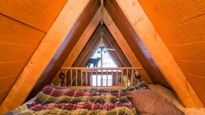 A triangular attic bedroom with a wooden sloped ceiling, a bed with a plaid blanket, and a window at the peak.