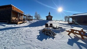 Un espace extérieur enneigé avec un bâtiment en bois, une petite construction dotée d’un toit en pente et une table de pique-nique.