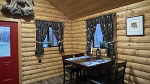 L’intérieur d’une cabane en rondins, avec une table à manger en bois, des chaises, deux fenêtres avec des rideaux, une porte rouge et une sculpture de loup en pierre accrochée au mur.