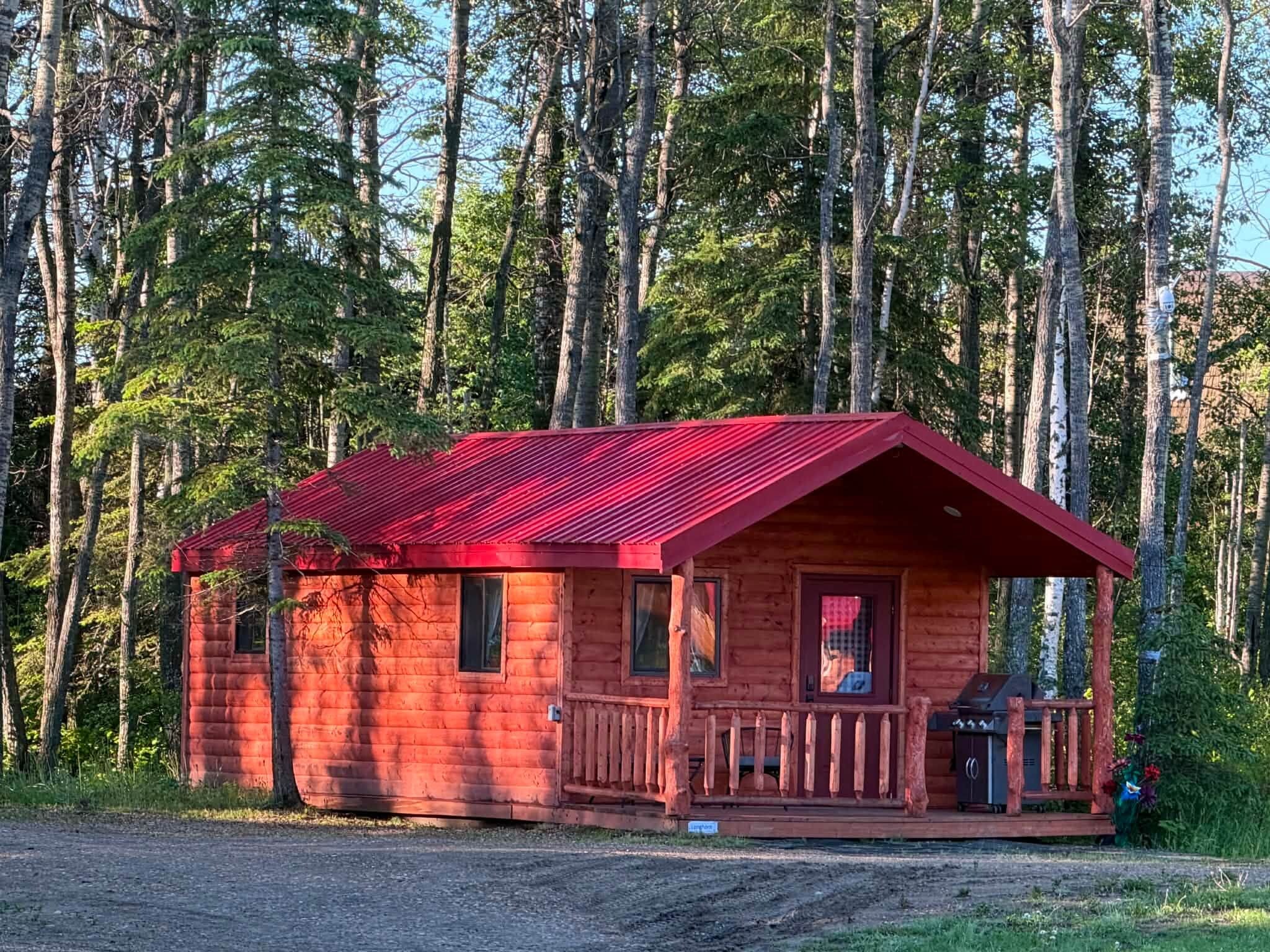 Una pequeña cabaña de madera con techo rojo, un porche y una chimenea, rodeada de un bosque.