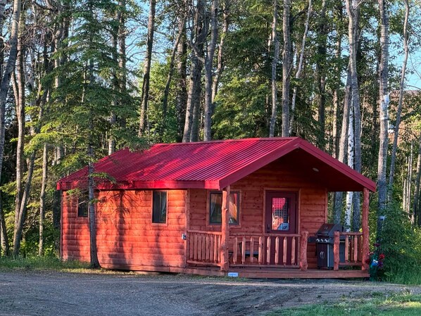 A small log cabin with a red roof, a porch, and a chimney, surrounded by a forest.