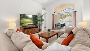 Un salon spacieux doté d’un grand téléviseur à écran plat, de canapés confortables, d’une table basse en bois et offrant une vue sur une piscine grâce à une porte vitrée.