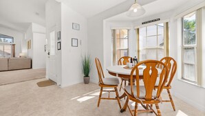 Une salle à manger lumineuse, avec une table ronde et des chaises en bois, une plante en pot, et une vue sur l’extérieur grâce à de grandes fenêtres.