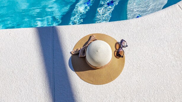 Un chapeau de paille, des lunettes de soleil et une serviette de plage sur une surface blanche texturée, près d’une piscine dont l’eau est d’un bleu limpide.