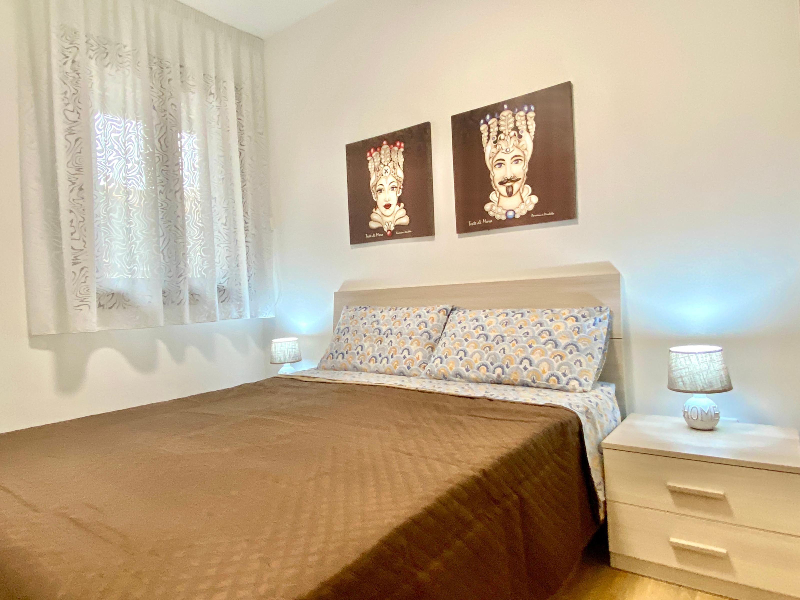 A bedroom with a bed, two wall-mounted lamps, a nightstand, and two framed pictures.