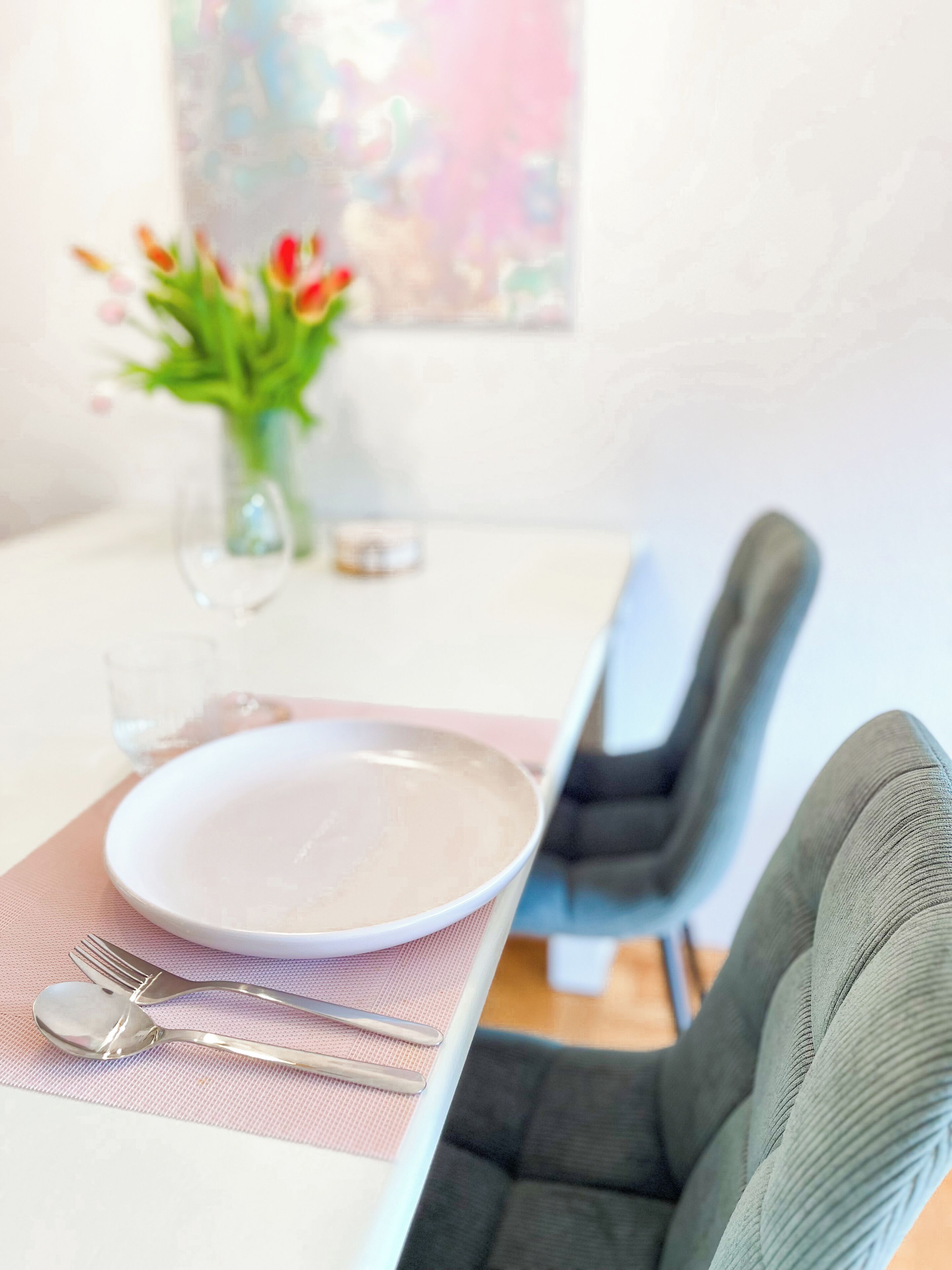 Une table à manger avec une assiette blanche, des couverts et un verre d’eau. Un vase rempli de fleurs est posé sur la table, et on aperçoit une chaise.