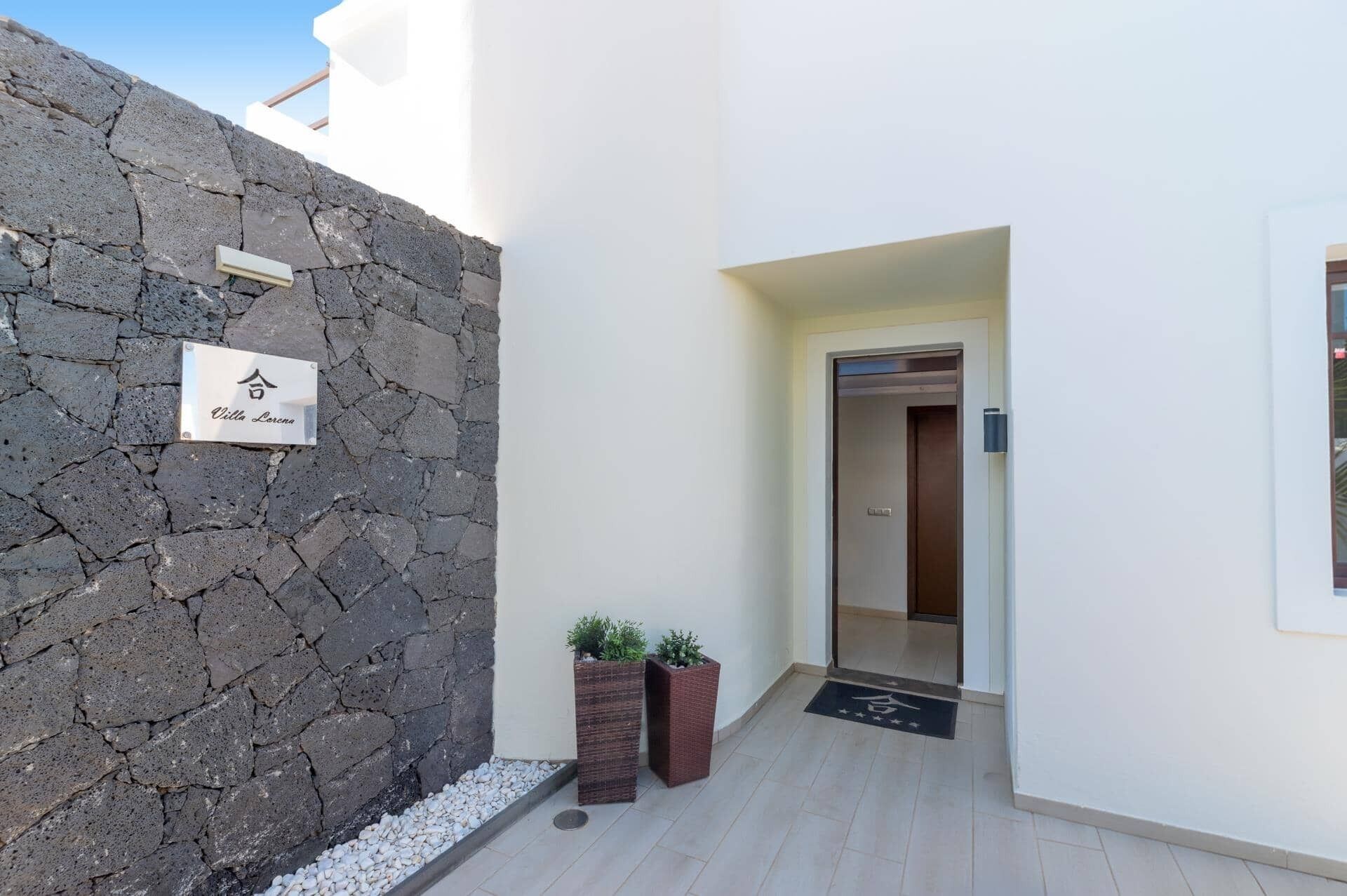 A narrow hallway with a stone wall on one side and a white wall on the other, leading to an elevator. There are two potted plants at the base of the white wall.