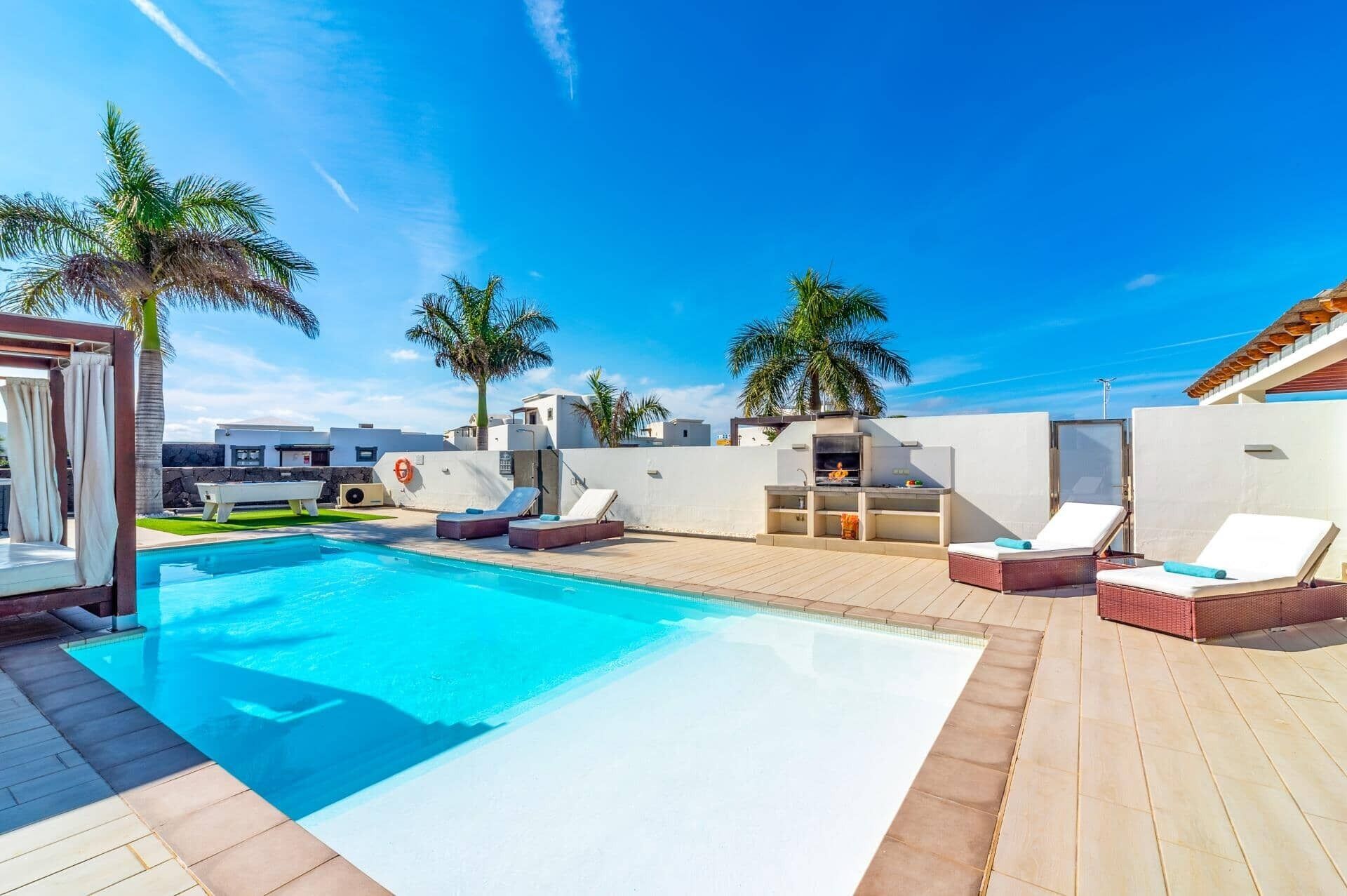 A swimming pool with lounge chairs and a barbecue area, surrounded by palm trees and modern buildings.