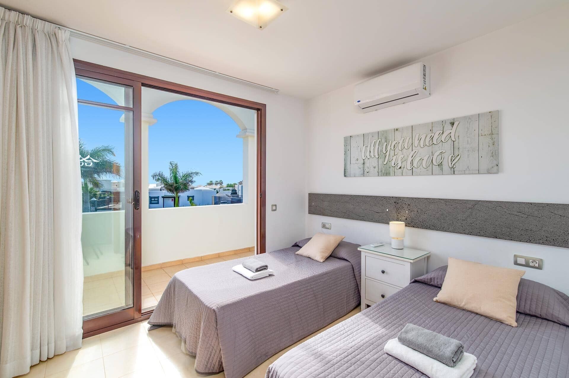 Quarto de hotel com duas camas, uma janela grande com cortinas, um ar condicionado e uma frase motivacional na parede.