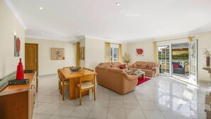 A spacious living area with a dining table and two sofas, a sliding glass door to an outdoor area, and a kitchen with a red decorative item on the counter. - Villa Caly by Villa Plus (Olhos de Água)