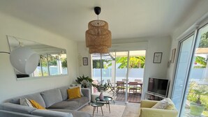 A modern living room with a grey sofa, yellow cushions, a small round table, and a yellow armchair. There is a large mirror on the wall, a hanging light fixture, and a TV on a stand.