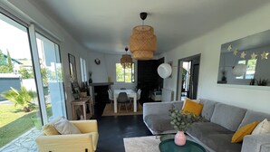 A modern living room with a sofa, armchair, and coffee table. There is a dining area with a table and chairs. The room has large windows and a view of the outdoors.