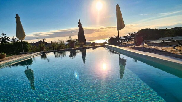 Ein klares Schwimmbecken mit Sonnenuntergangsblick, umgeben von Sonnenschirmen und einer Sandfläche.
