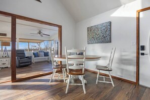 A dining area with a round wooden table and white chairs, a ceiling fan, and a view of a living room with a sofa and a window.
