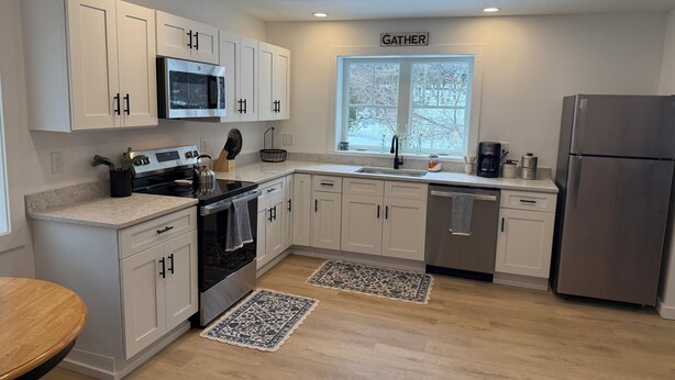 A modern kitchen with white cabinets, stainless steel appliances, a window with a view, and a 'GATHER' sign above the window.
