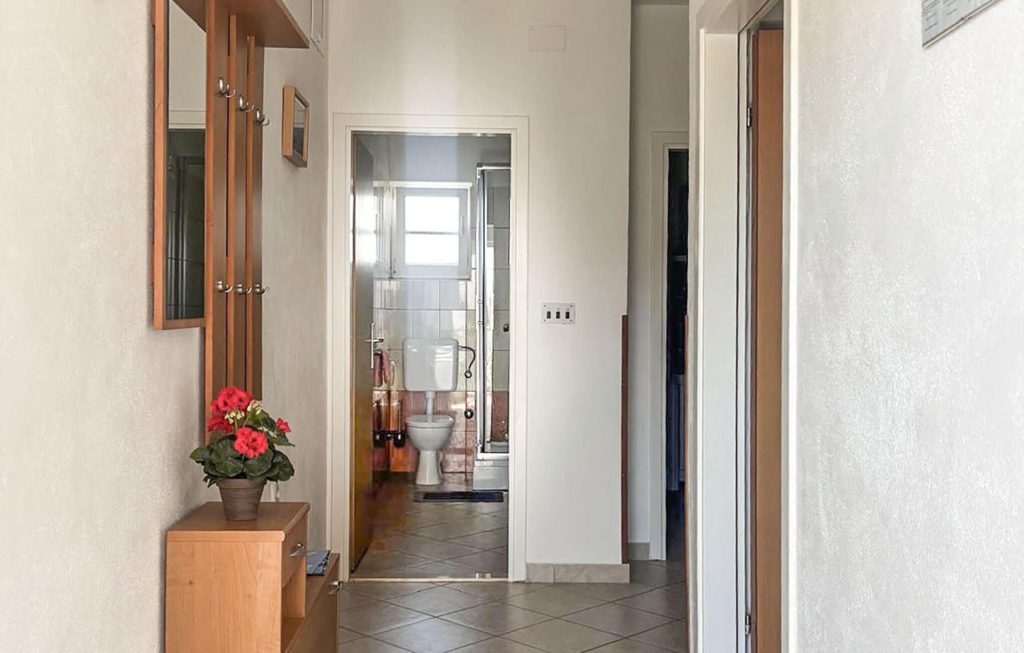 Ein Flur, der zu einem Badezimmer mit Toilette, Waschbecken und Dusche führt. Ein Holzschrank mit einem Topfpflanzen darauf ist ebenfalls zu sehen.