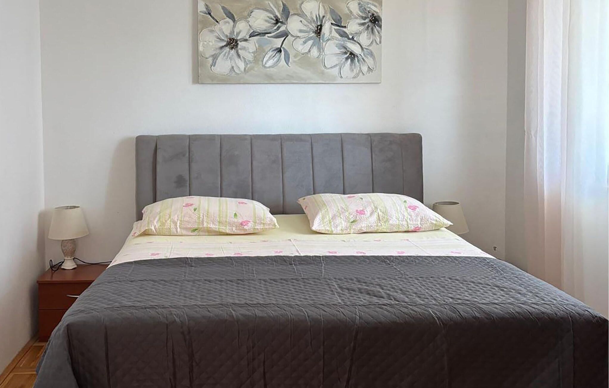 Ein Schlafzimmer mit grauem, gepolsterten Kopfteil, zwei Kissen mit Blumenmuster, einem Bett mit grauer Tagesdecke, einem Nachttisch mit Lampe und einem Wandbild von Blumen.