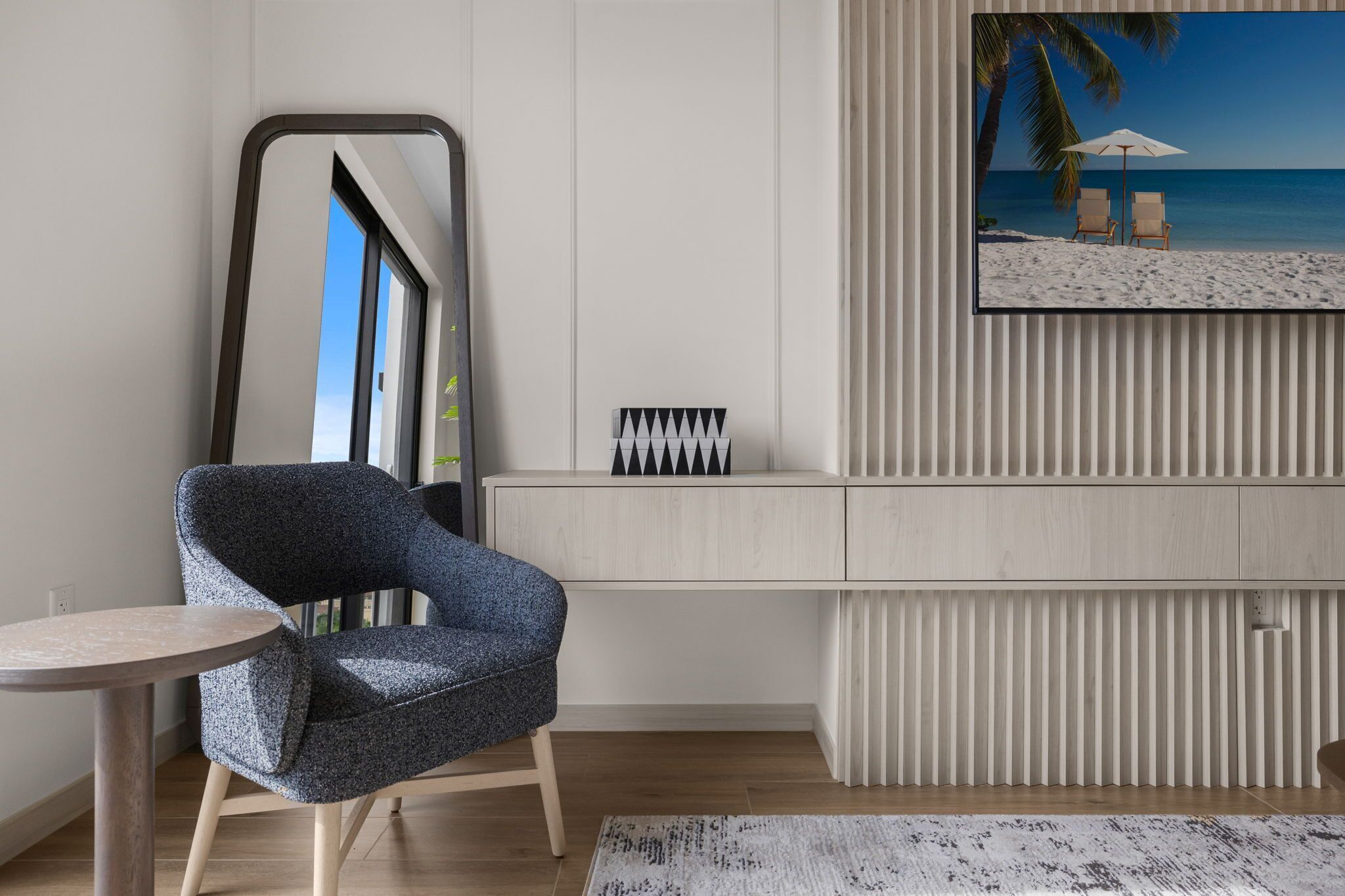 A modern room with a large window, a blue armchair, a small round table, a framed picture of a beach, and a white cabinet with a geometric design.