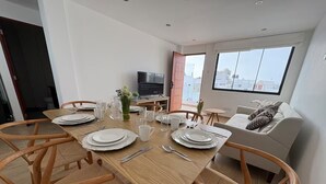 Een eethoek met een houten tafel gedekt voor vier personen, omringd door rieten stoelen. Een televisie is aan de muur bevestigd, en een bank is op de achtergrond te zien.