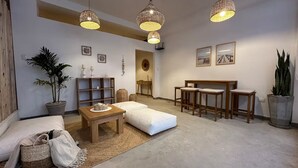 Een moderne woonkamer met een bank, een salontafel en een eethoek met krukken. Er staan rieten lampen, een potplant en er hangen fotolijstjes aan de muren.
