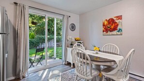 Un coin repas avec une table et des chaises, un réfrigérateur et une vue sur un jardin, accessible par une porte-fenêtre coulissante.