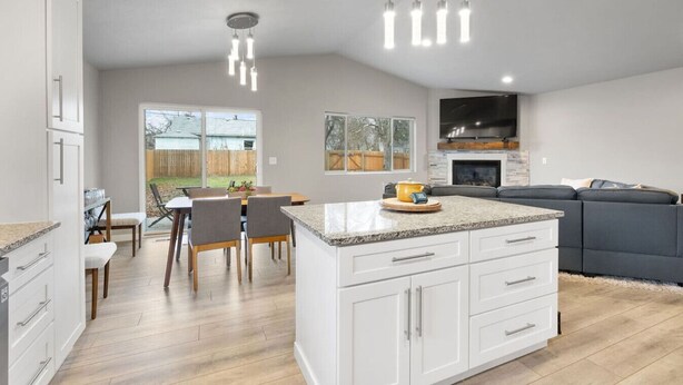 A modern kitchen with white cabinetry, a granite countertop island, and a dining area with a table and chairs. - Near Perry, Near Perfect (Spokane)