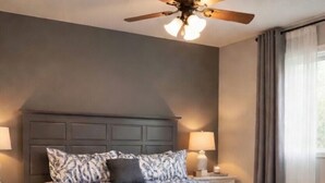Ein Schlafzimmer mit Bett, Nachttischen, Deckenventilator, Fenster mit Vorhängen und einer dunkelbraunen Bank.