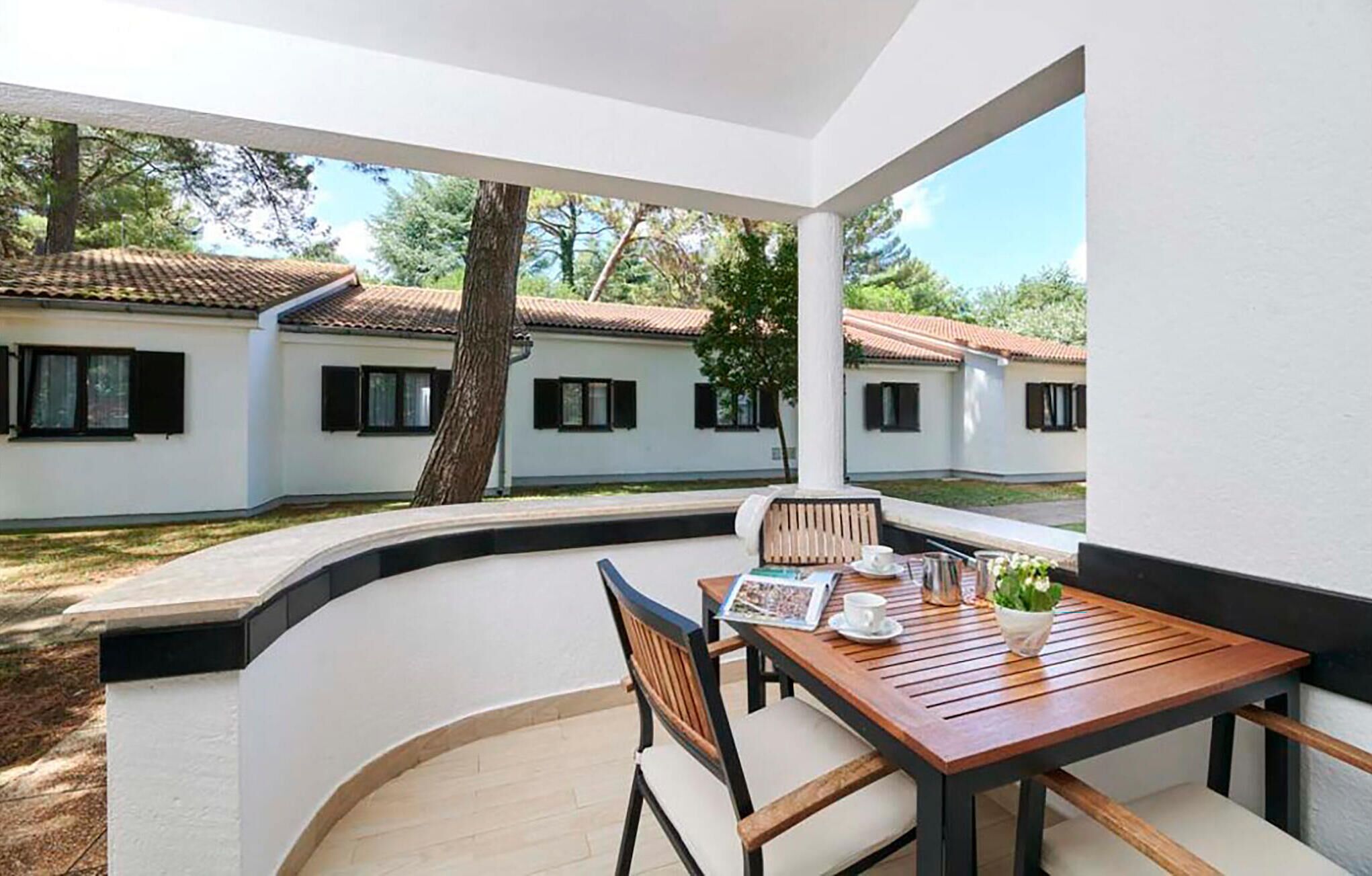 A patio with a wooden table and chairs, a view of white buildings with red roofs, and a tree in the background.