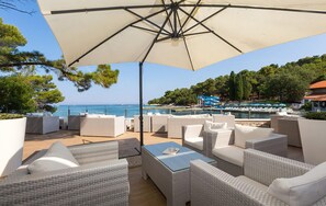 Un patio frente al mar con mobiliario de mimbre, un paraguas grande y vistas al mar y a una zona de piscina.
