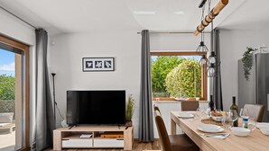 Un espace repas moderne avec une table en bois dressée pour deux personnes, un téléviseur à écran plat et une vue sur la verdure environnante.
