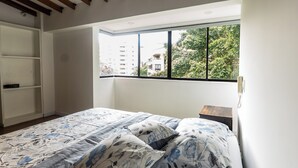 A bedroom with a large bed, a window with a view of buildings and trees, and built-in shelving. - Duplex en el Primer Parque de laureles (Medellín)