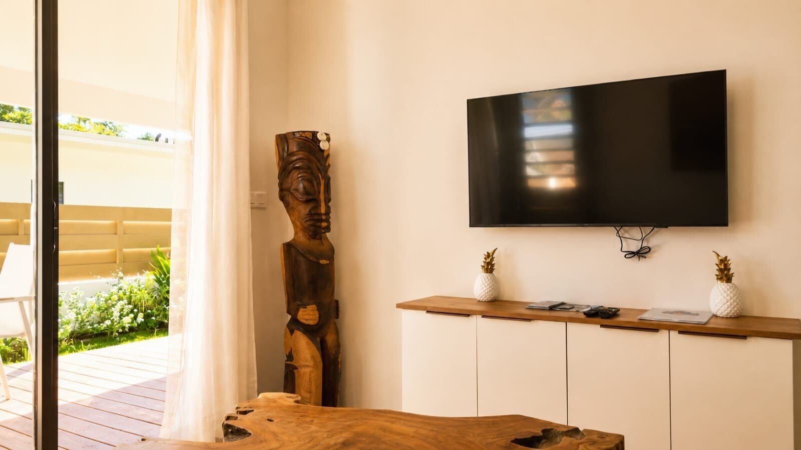 A room with a wooden sculpture, a TV mounted on the wall, and a wooden table with a white vase.