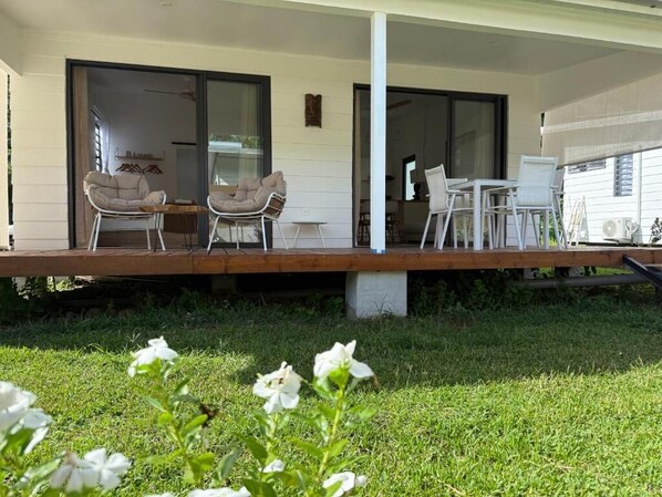 Eine überdachte Terrasse mit Sitzgelegenheiten im Freien, einer Betonstütze und Blick auf das Haus durch Glastüren.