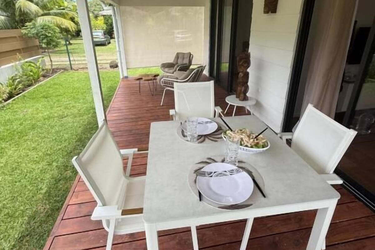 An outdoor dining area with a table set for two, surrounded by white chairs, on a wooden deck with a grassy lawn and palm trees in the background.
