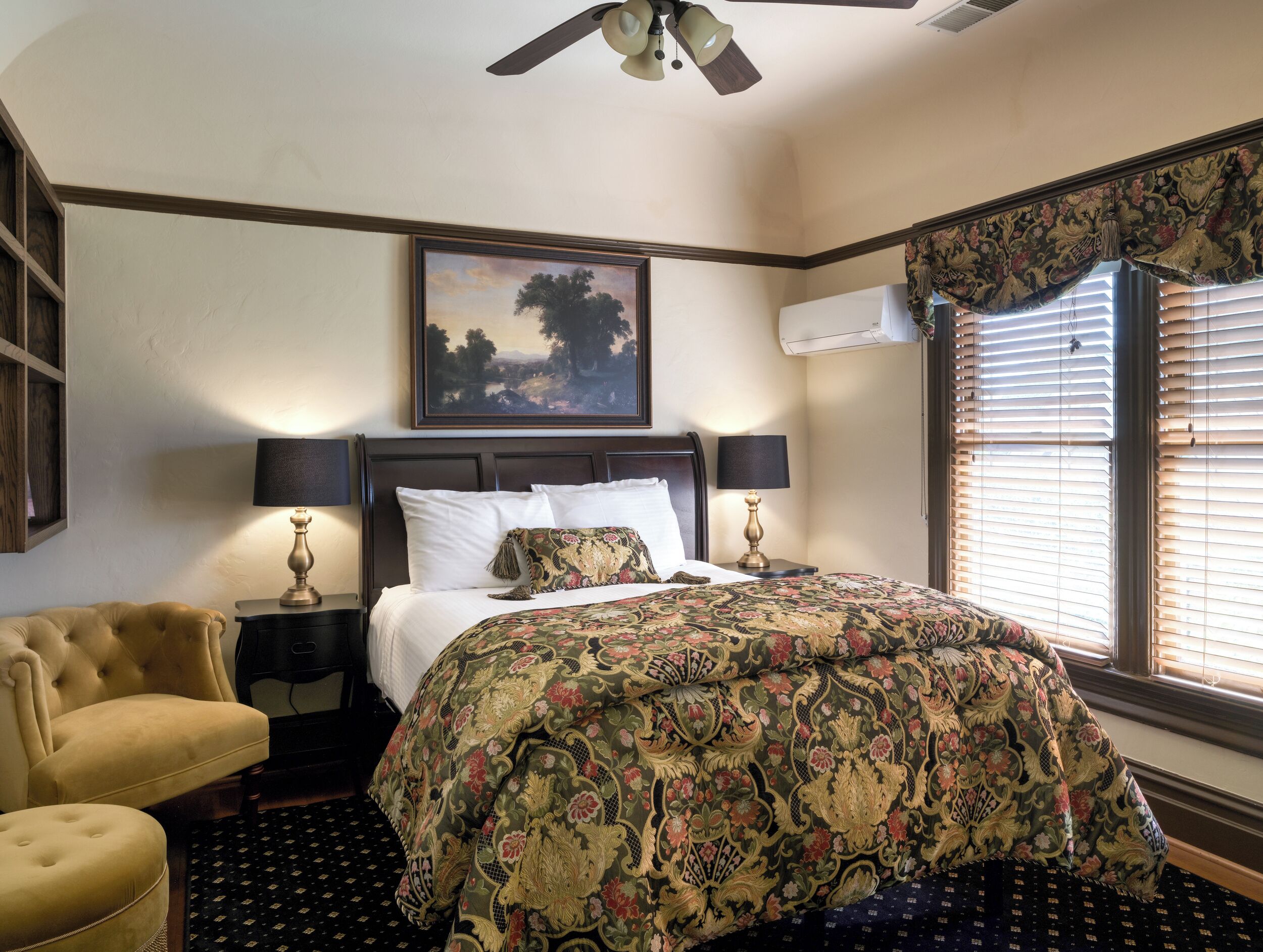 A hotel room with a large bed, two bedside tables with lamps, a chair, a painting on the wall, and a window with blinds.