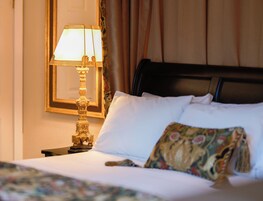 Cama bem arrumada com lençóis brancos e almofada decorativa, abajur dourado em uma mesa de cabeceira e espelho emoldurado na parede.
