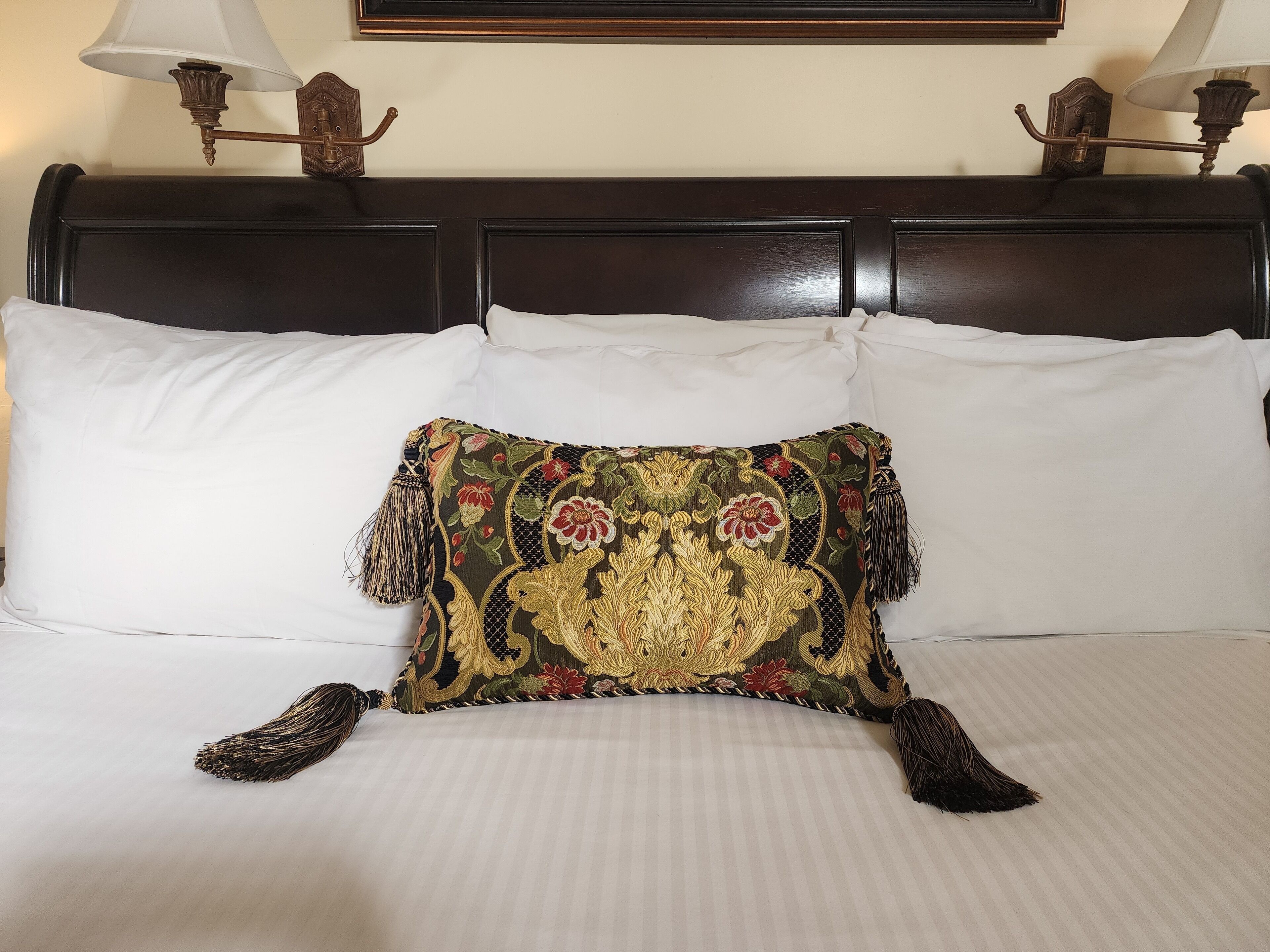 Una cama con sábanas blancas, un cojín decorativo bordado y dos lámparas fijadas a la pared.