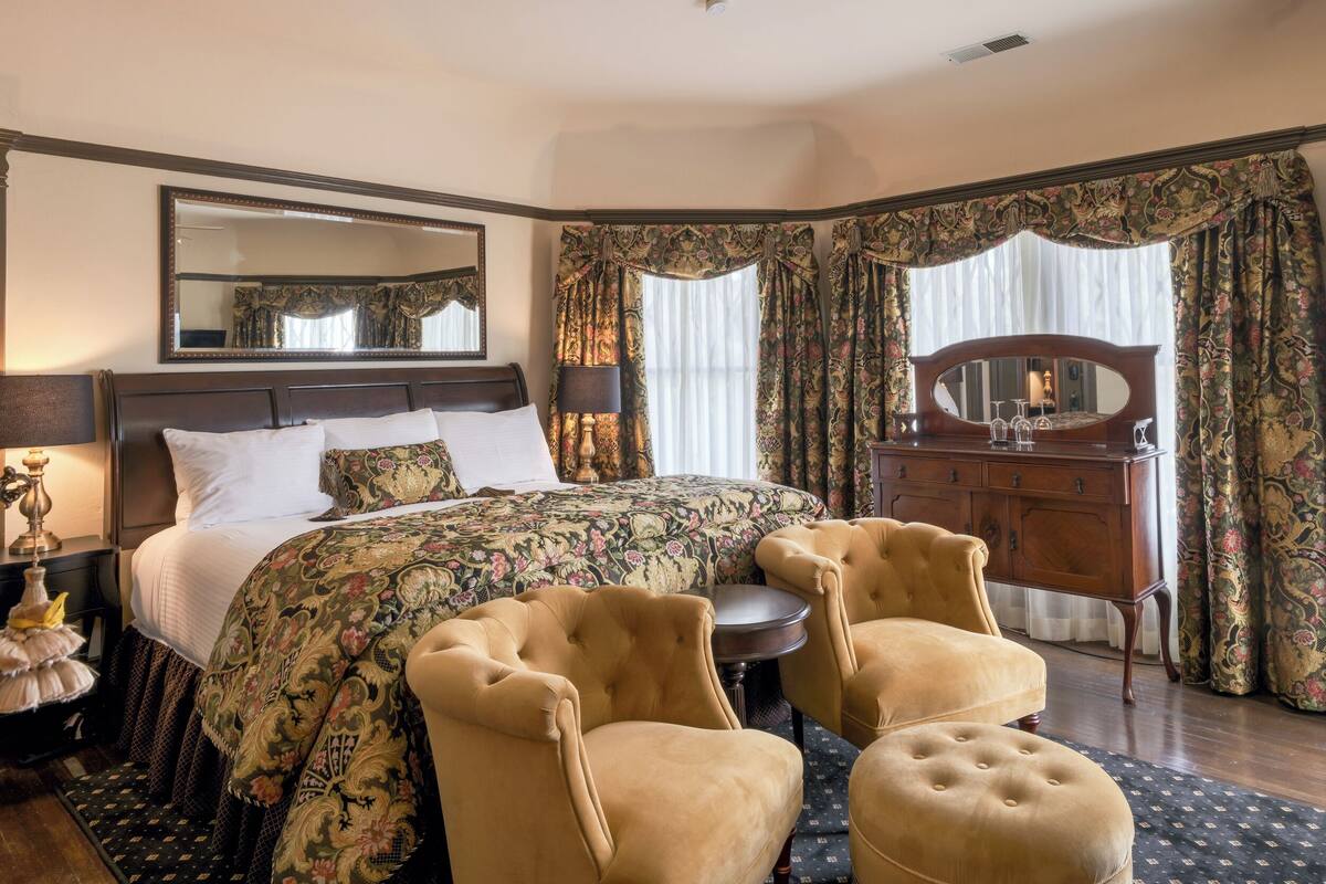 Une chambre d’hôtel avec un grand lit, une commode en bois, deux fauteuils, une table ronde et un miroir.