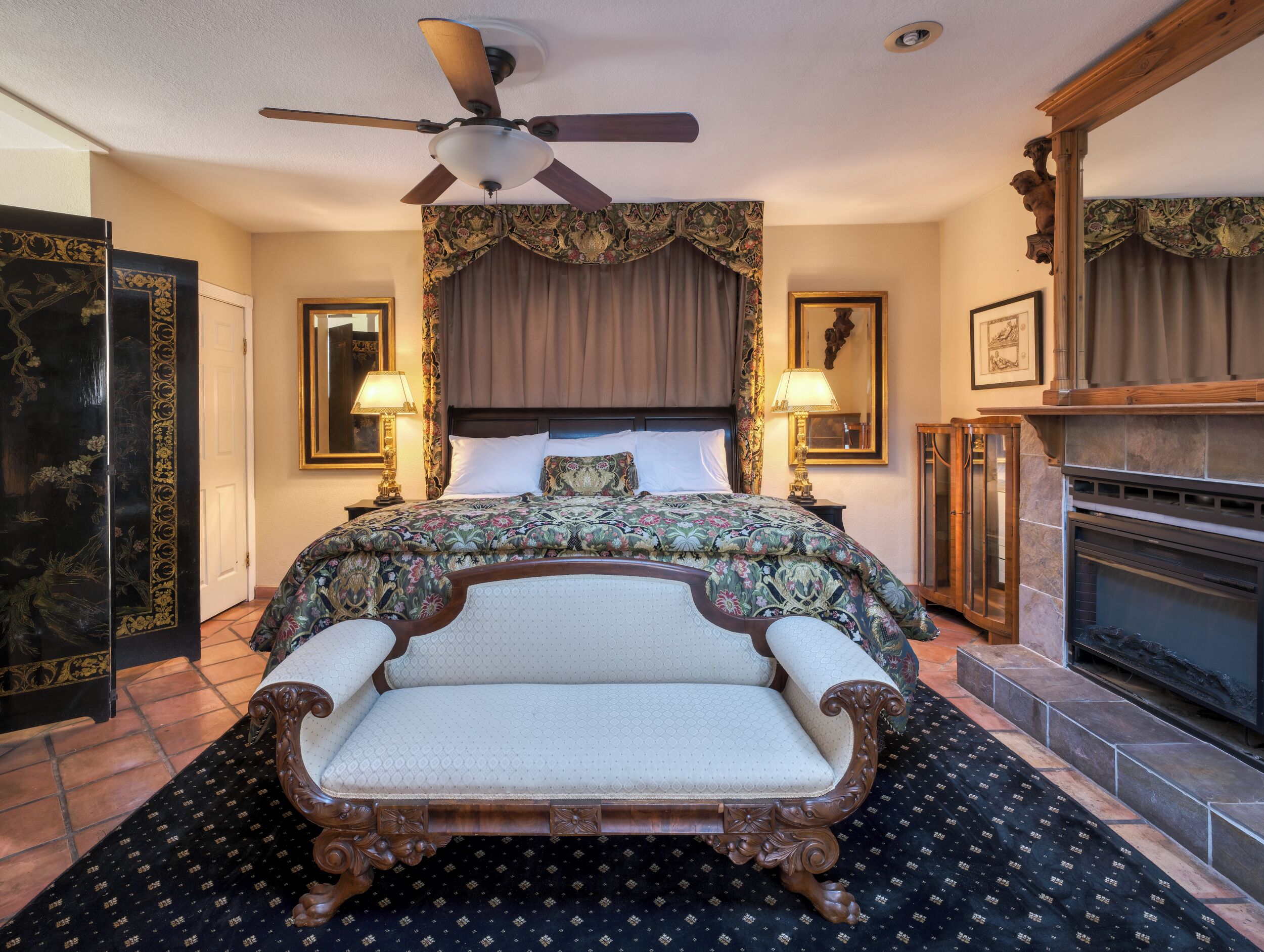 A hotel room with a large bed, a fireplace, a ceiling fan, and a large mirror.