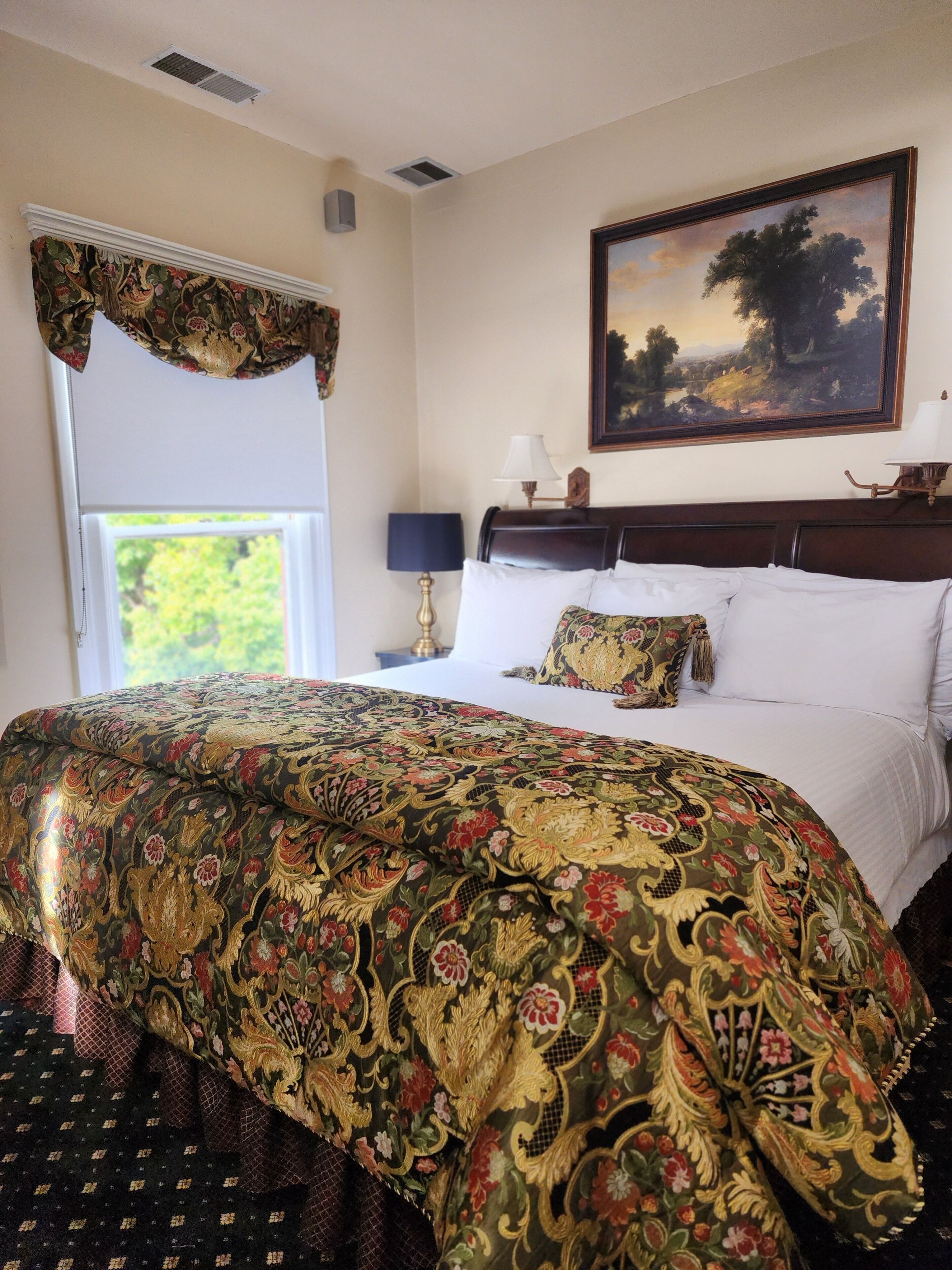 A hotel room with a large bed, a painting on the wall, and a window with a view of greenery.