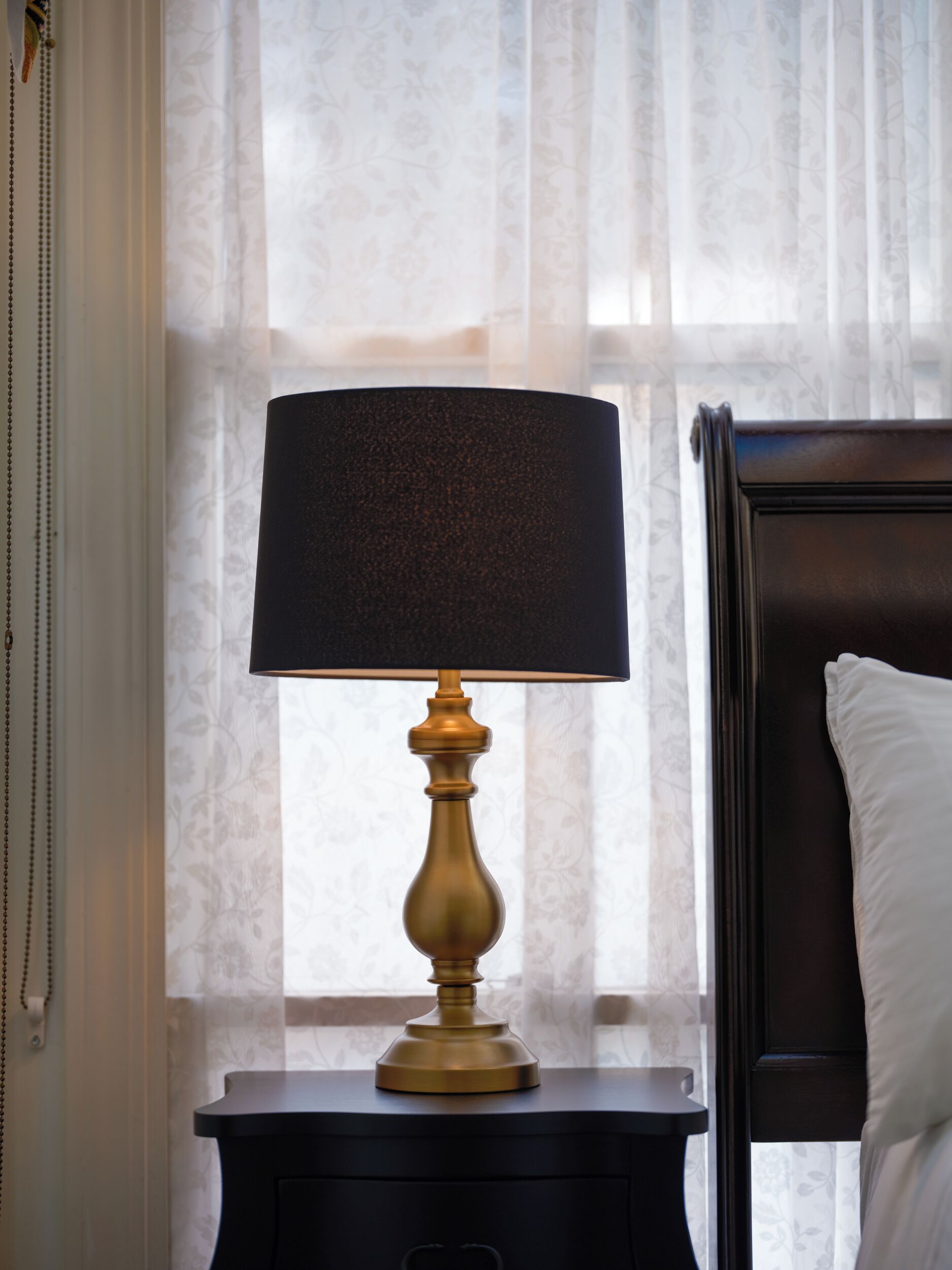 A bedside table with a lamp, a bed with a headboard, and a window with sheer curtains.