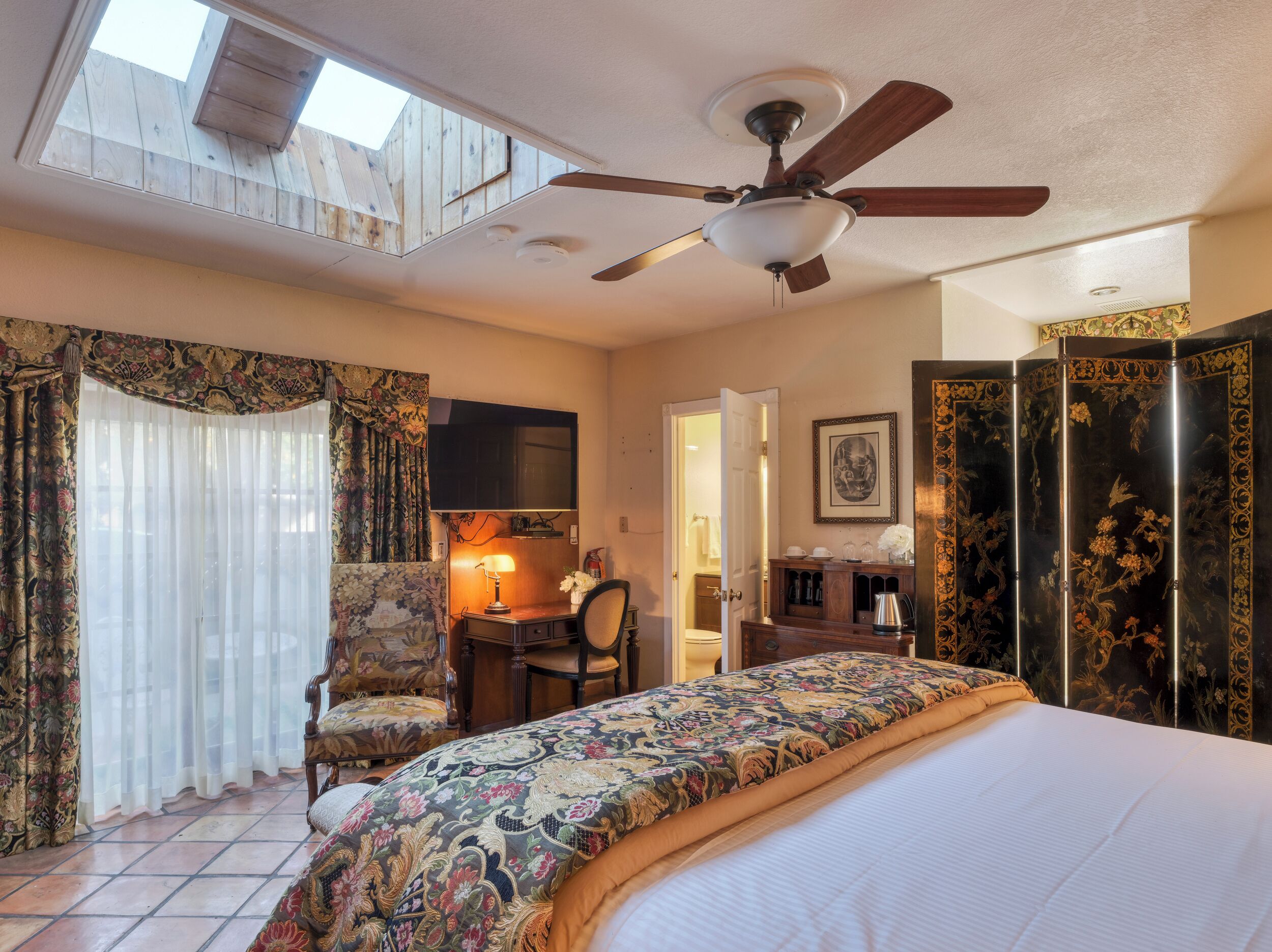 A hotel room with a bed, a desk, a chair, a ceiling fan, and a decorative screen.