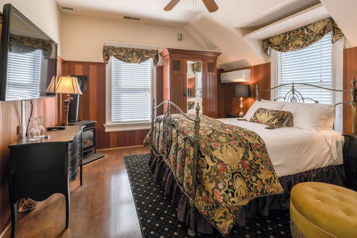 Une chambre à coucher comprenant un lit, un bureau, une chaise, une lampe, un ventilateur de plafond et une fenêtre avec des rideaux.