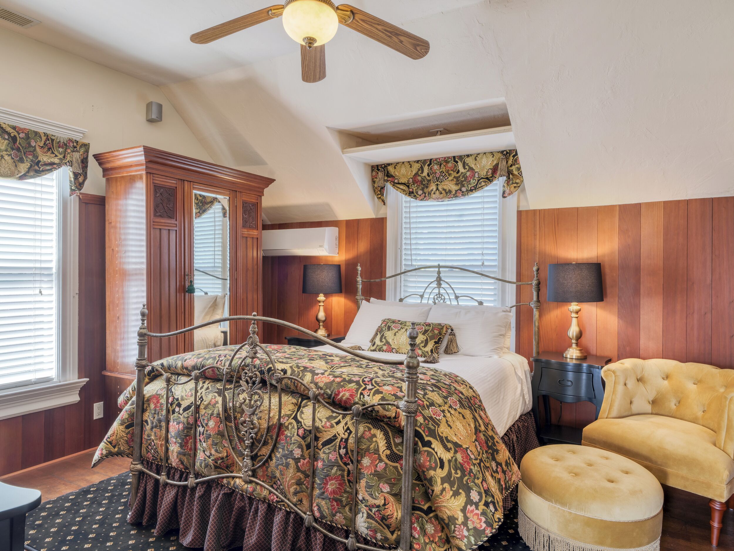 A bedroom with a wooden bed frame, a ceiling fan, a large window with blinds, and a wooden wardrobe.