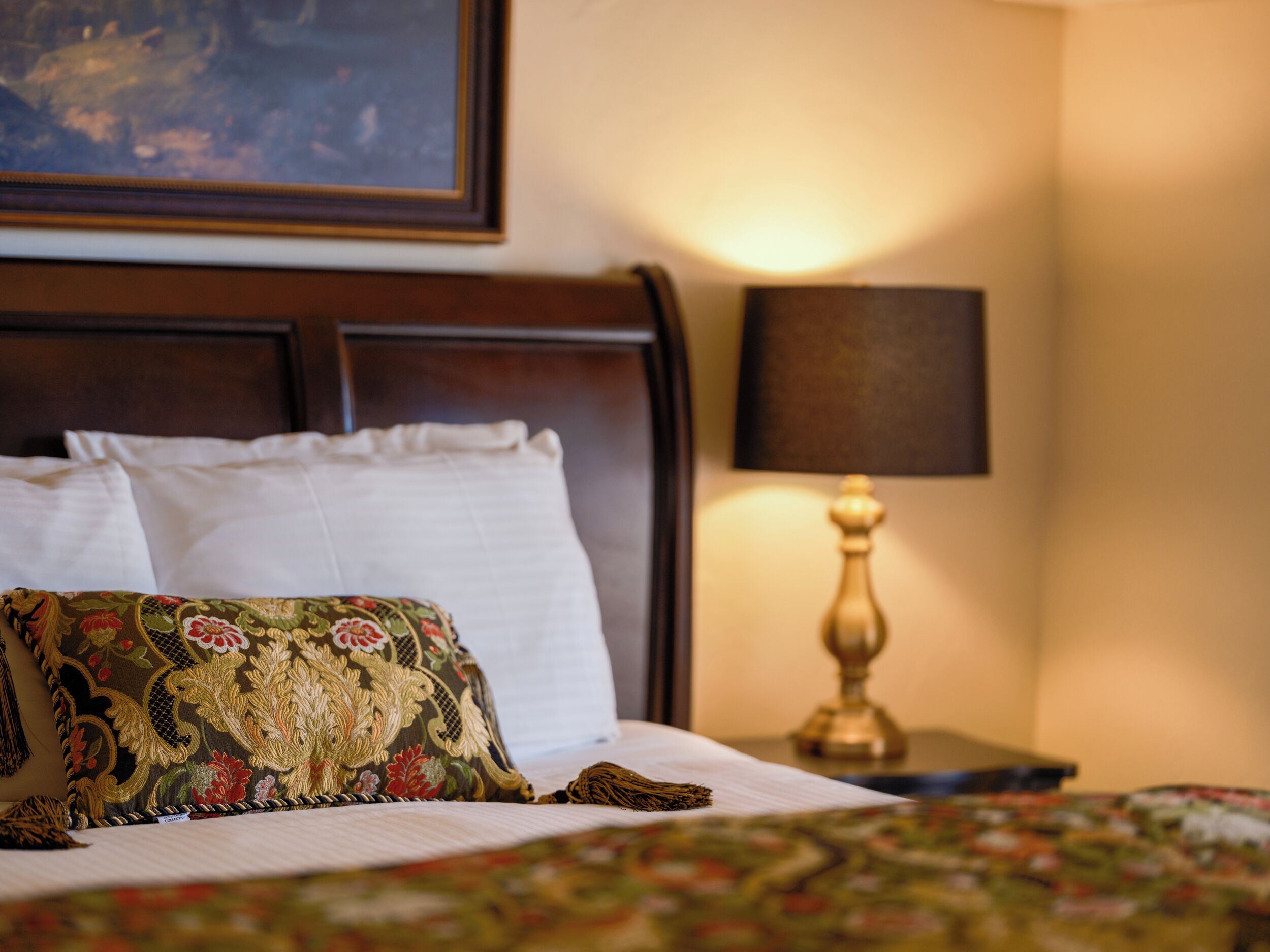 Una cama bien hecha con almohadones florales, un cabecero de madera y una mesita de noche con una lámpara.