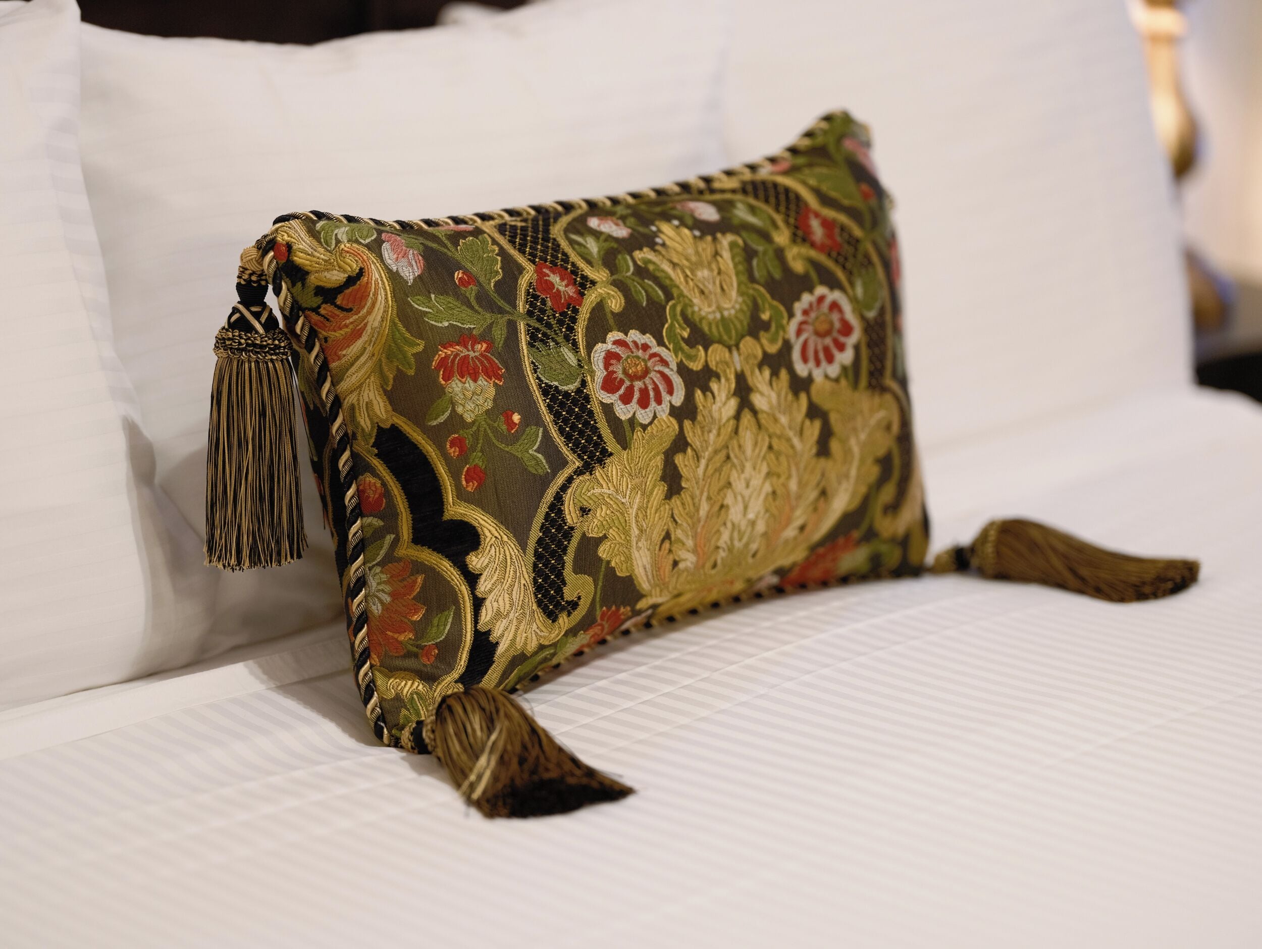 Un coussin orné d’un motif floral et de glands, posé sur un lit recouvert de draps blancs.
