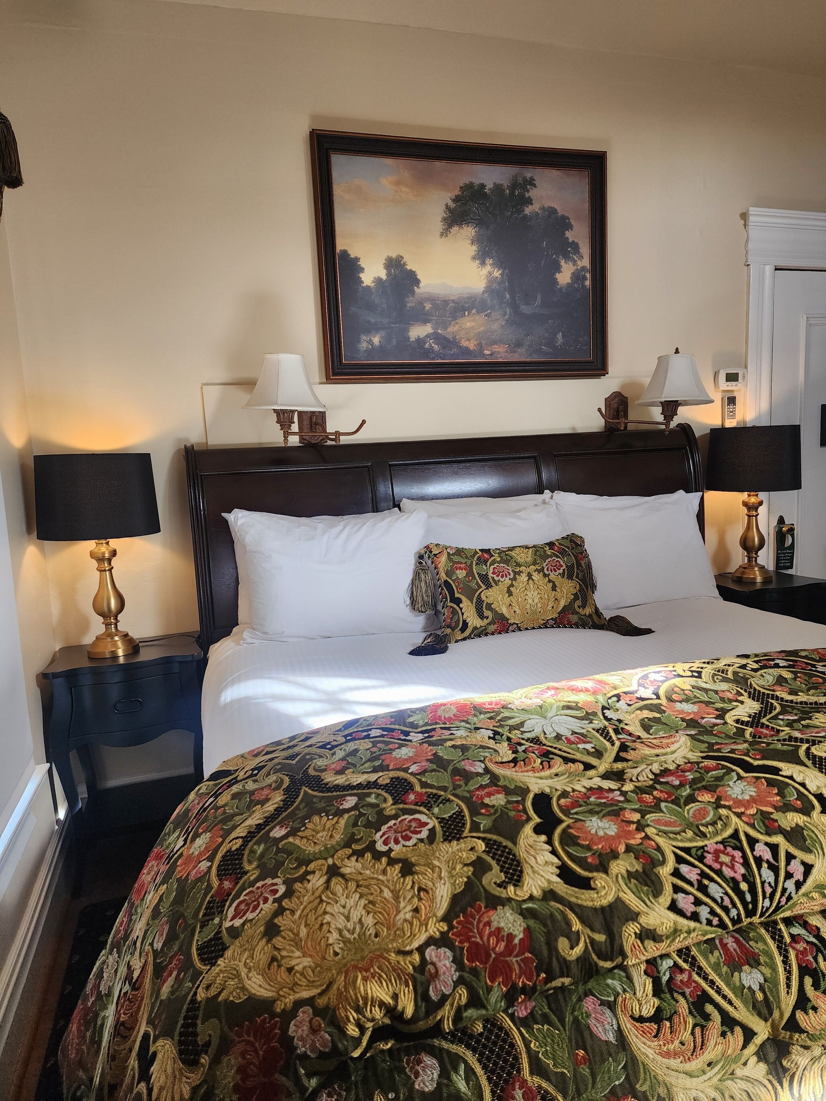 A neatly made bed with a floral bedspread, two bedside lamps, a nightstand, and a painting above the bed.