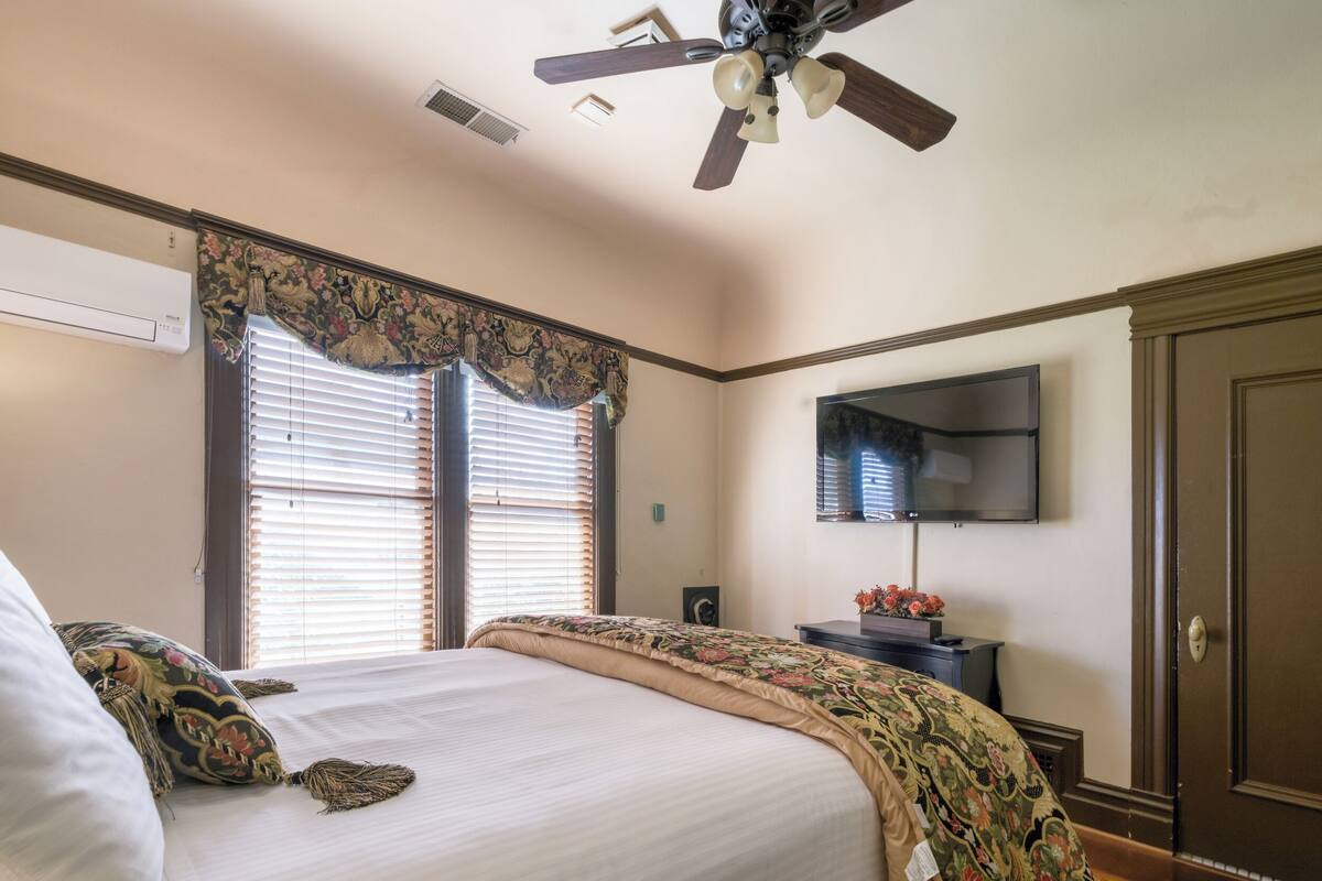 Une chambre à coucher avec un lit, un ventilateur de plafond, un téléviseur fixé au mur et une porte.